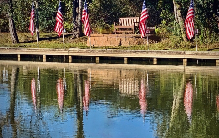 Five American flags and a bench on a dock by a lake with their reflections in the water on a sunny day.