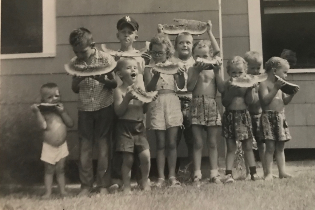 Black-and-white photo of eight children standing outside eating large slices of watermelon in front of a house.