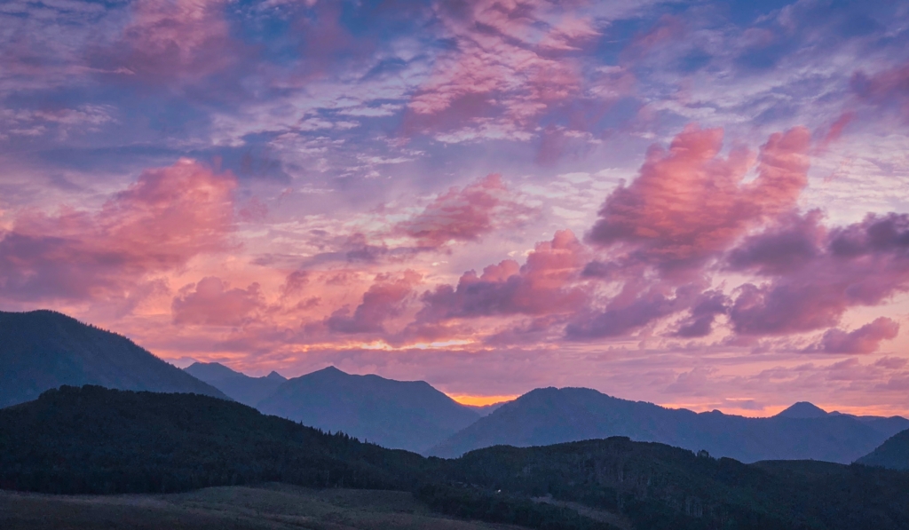 sunrise over the mountains with pink, blue, and gray clouds with a hint of the rising sun at the peak of the mountains