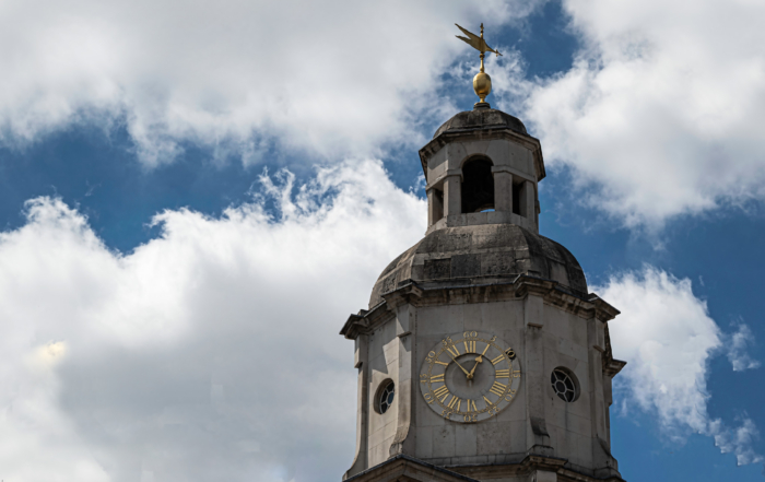 Clock tower with Roman numerals and a weather vane under a partly cloudy blue sky.