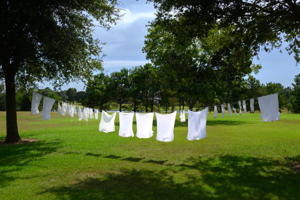 White clothes hanging on a clothesline between trees in a sunny, green open field under a partly cloudy sky.