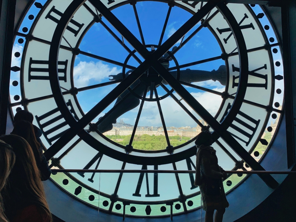 View from inside a large clock face with Roman numerals, showing a cityscape and people silhouetted against a bright blue sky.