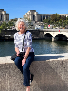 Smiling woman in a striped shirt sits beside the Seine in Paris, with a stone bridge, grand historic buildings, trees, and blue sky behind her.