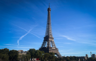 Eiffel Tower rises above Paris trees and the Seine River beneath a vivid blue sky with wispy clouds, creating an iconic, serene scene.