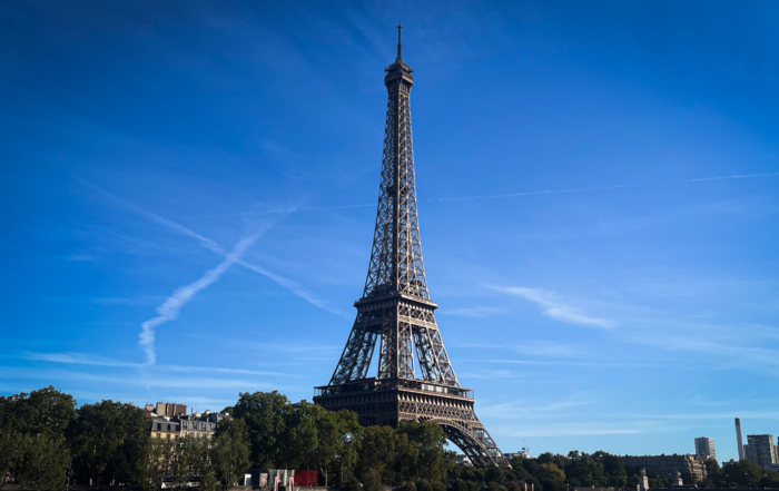 Eiffel Tower rises above Paris trees and the Seine River beneath a vivid blue sky with wispy clouds, creating an iconic, serene scene.