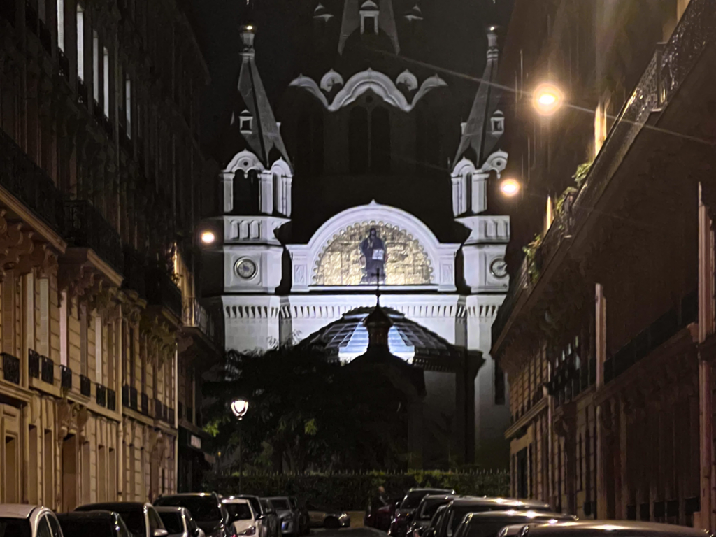 Illuminated ornate cathedral façade viewed at night from a narrow street lined with historic buildings and parked cars.