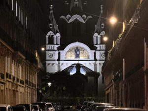 Illuminated ornate cathedral façade viewed at night from a narrow street lined with historic buildings and parked cars.