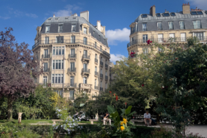 Sunlit Parisian park with flowering gardens and seated visitors, framed by elegant cream Haussmann buildings under a vivid blue sky.