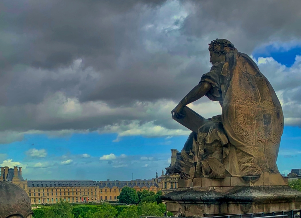Seated stone statue of a robed figure overlooking formal gardens and historic Parisian buildings beneath a dramatic cloudy sky.