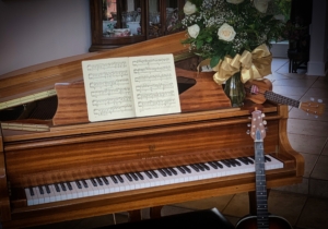 Open sheet music rests on an upright piano beside white roses and a guitar.