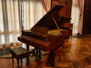 Open grand piano with raised lid and bench in an elegant room with curtains and polished wood floors.