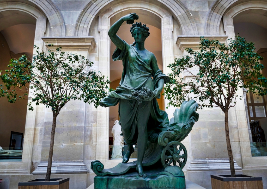 Bronze statue of Aurora, the Goddess of Dawn in the Louvre, a seated woman with raised arm, surrounded by leafy trees in a stone-arched courtyard.